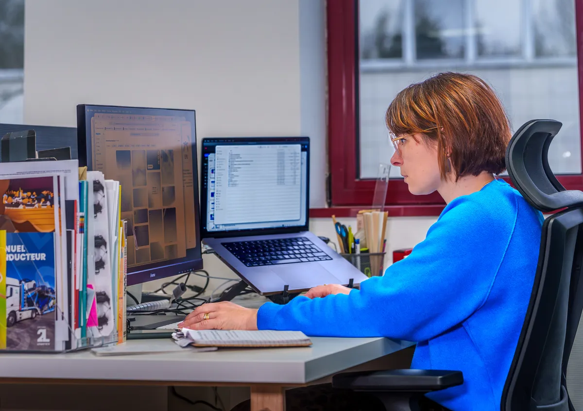 Graphiste EcloLINK à son poste de travail double écran, entourée de réalisations print de l'agence